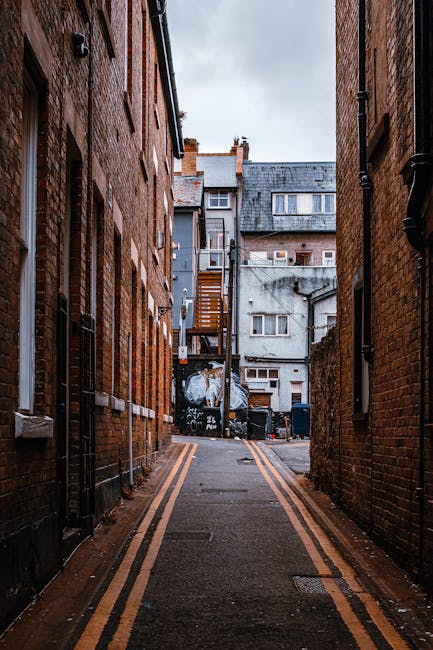 A narrow urban alleyway bordered by tall brick buildings on either side, with a paved surface marked by double yellow lines along its edges. In the background, there are residential structures with va