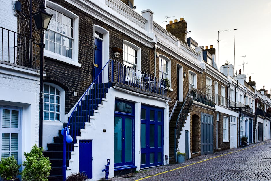 A narrow urban alleyway bordered by tall brick buildings on either side, with a paved surface marked by double yellow lines along its edges. In the background, there are residential structures with va