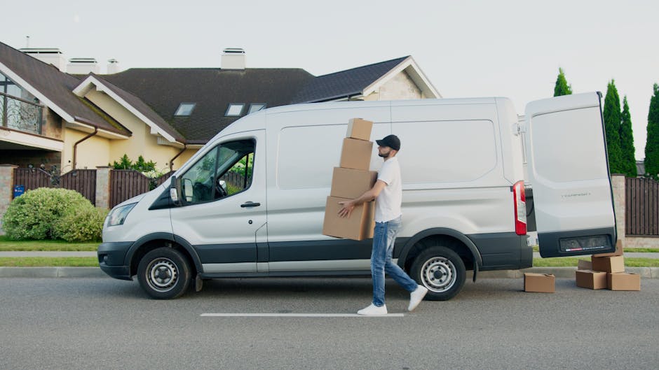 A man wearing a white t-shirt, jeans, and a cap is engaged in home relocation activities, carrying three stacked cardboard boxes towards a white panel van parked on a residential street. The van, used