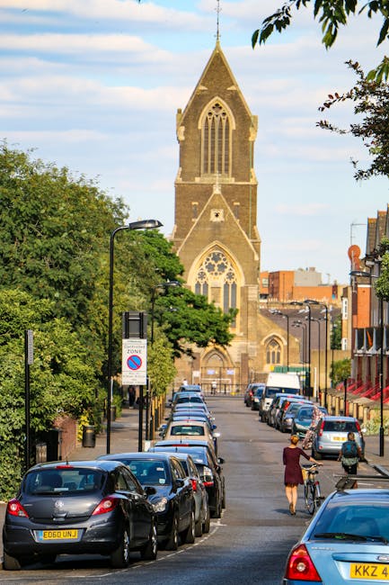 A man wearing a white t-shirt, jeans, and a cap is engaged in home relocation activities, carrying three stacked cardboard boxes towards a white panel van parked on a residential street. The van, used