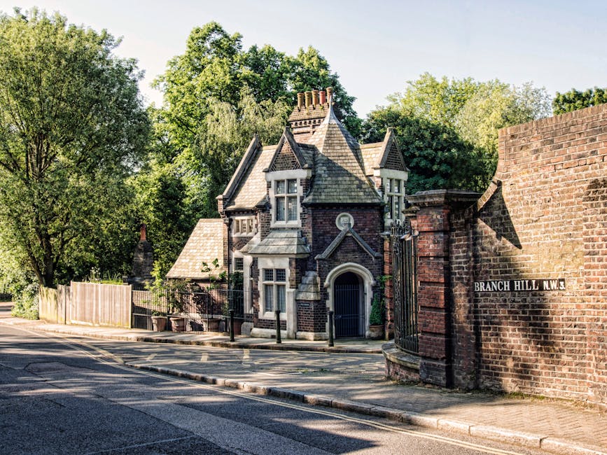 A Victorian-style house situated on a residential street, featuring a steeply pitched roof with multiple gables, ornate brickwork, and bay windows. The property is enclosed by a black metal fence with a small gate, and there is a paved sidewalk along the street. Tall, leafy trees surround the house, casting shadows on the pavement. Adjacent to the house, a brick wall displays a street sign reading 'Branch Hill N.W.3'. The scene captures a clear, sunny day with natural light illuminating the building and surrounding greenery, representing a typical location for house removals or home relocation services provided by Man and Van Dulwich. The environment appears calm and well-maintained, with the aesthetics of the street highlighting a classic British residential area suitable for furniture transport and packing and moving operations.